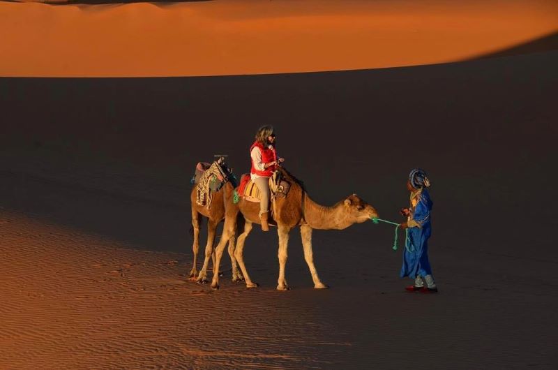 Woman riding a camel in the sunset while the guide is helping them both out, the camel and the woman 